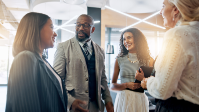 Four colleagues smile and chat in a bright, modern office space, standing in a loose circle.
