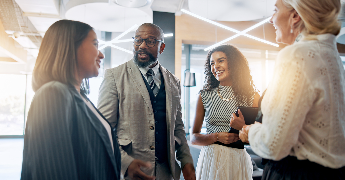 Four colleagues smile and chat in a bright, modern office space, standing in a loose circle.