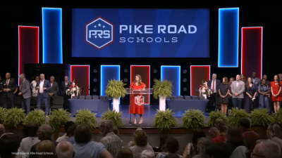 Speaker in a red dress delivers a speech at a podium on stage, with Pike Road Schools branding and blue-red backlit panels behind her, flanked by a line of people and potted ferns, audience in foreground.
