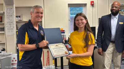 Man in a dark blue and yellow polo presents certificate to a young woman in a yellow shirt; a suited man watches in a room with shelves and a window in the background.