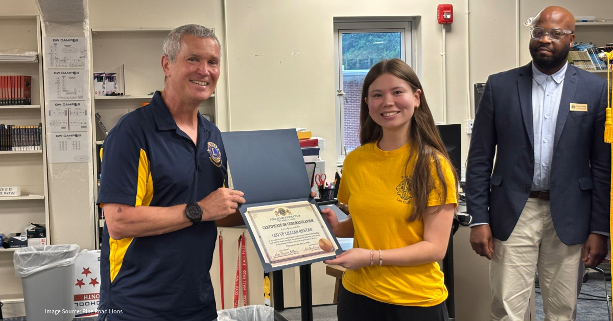 Man in a dark blue and yellow polo presents certificate to a young woman in a yellow shirt; a suited man watches in a room with shelves and a window in the background.
