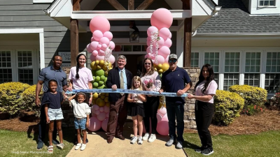 Group of adults and kids pose with a ribbon-cutting ceremony in front of a house, pink balloon arches behind them and a 'Welcome Home' ribbon.