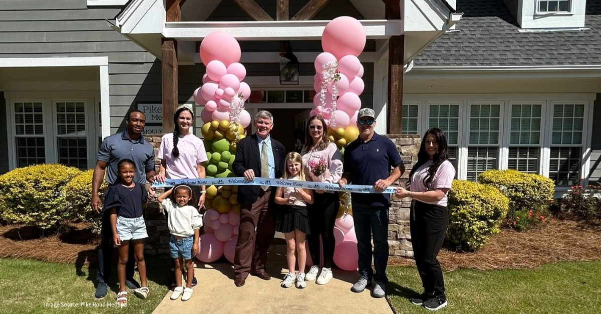 Group of adults and kids pose with a ribbon-cutting ceremony in front of a house, pink balloon arches behind them and a 'Welcome Home' ribbon.